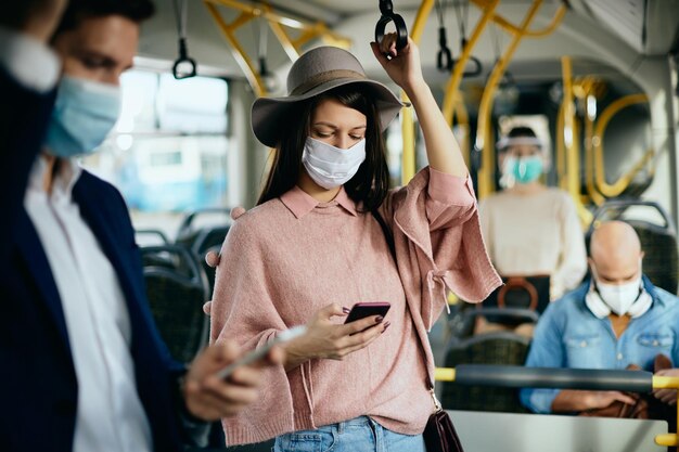 Donna con maschera facciale protettiva che invia messaggi di testo sul telefono cellulare durante il pendolarismo in autobus