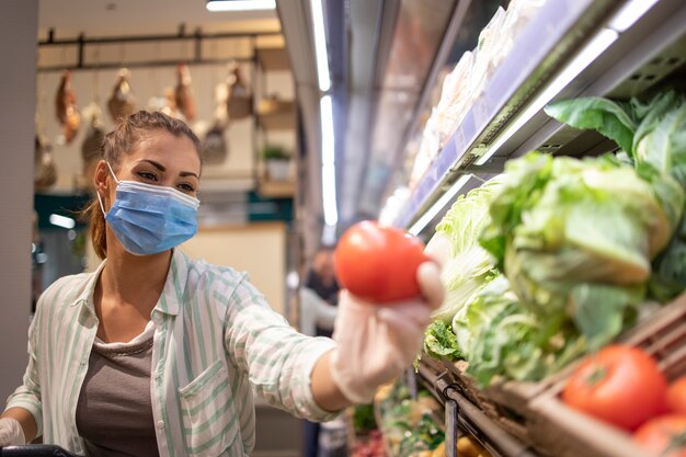 Donna con maschera igienica e guanti di gomma e carrello della spesa in drogheria acquistare verdure durante il virus corona e prepararsi per una quarantena pandemica