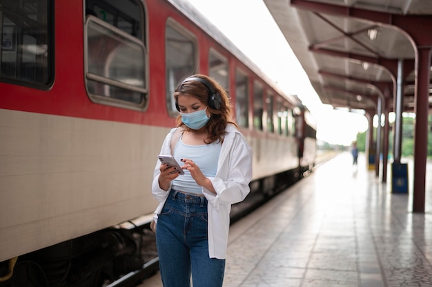 Donna con maschera medica che utilizza smartphone alla stazione ferroviaria pubblica