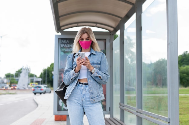 Donna con maschera medica che utilizza smartphone mentre aspetta l'autobus pubblico