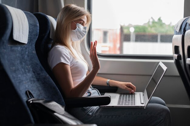 Donna con maschera medica che viaggia in treno pubblico e fa una videochiamata sul laptop