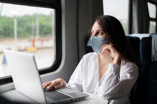 Donna con maschera medica che viaggia in treno pubblico e usa il laptop