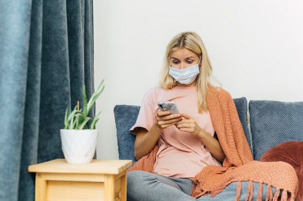 Donna con maschera medica e smartphone a casa durante la pandemia