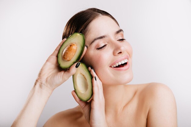 Donna con spalle nude sorrisi carini in posa con avocado utile per la pelle su sfondo bianco