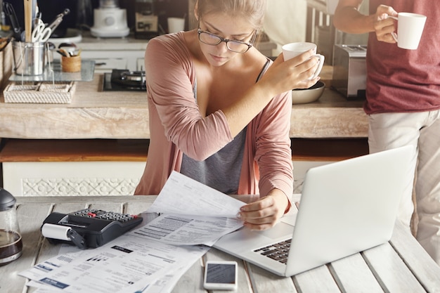 Donna concentrata vestita casualmente calcolando le bollette, seduto al tavolo della cucina con laptop, calcolatrice, documenti e cellulare, tenendo la tazza bianca e passandola a suo marito