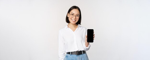 Donna coreana sorridente che mostra lo schermo dello smartphone che mostra l'applicazione mobile in piedi sopra il bianco