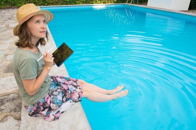 Donna creativa ispirata in cappello di estate che si siede a bordo piscina