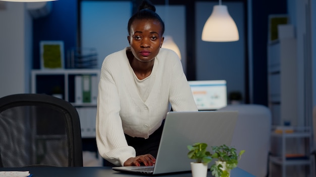 Donna d'affari africana in piedi vicino alla scrivania guardando la telecamera dopo aver letto la posta sul laptop che lavora in una start-up a tarda notte. Dipendente concentrato che utilizza la tecnologia wireless di rete facendo gli straordinari