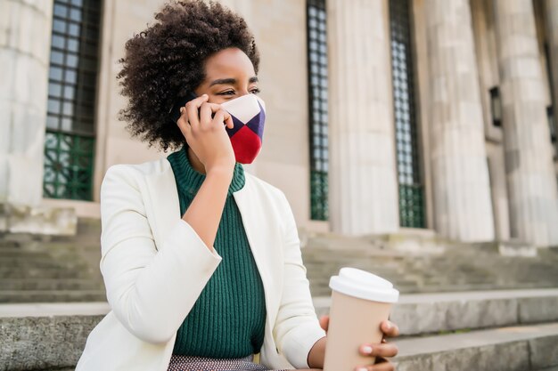 Donna d'affari afro che indossa la maschera protettiva e parla al telefono mentre era seduto sulle scale all'aperto in strada. Business e concetto urbano.