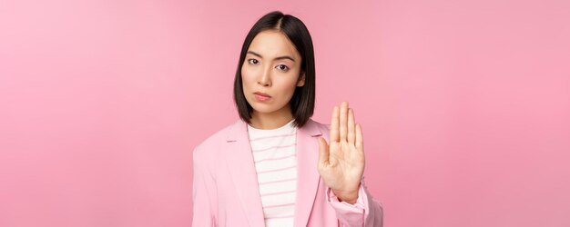 Donna d'affari asiatica con espressione faccia preoccupata seria che mostra tabù stop motion proibire il gesto disapprovare smth cattivo in piedi su sfondo rosa