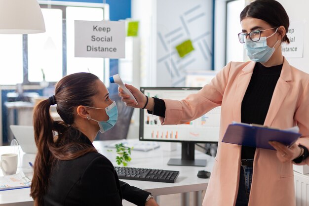 Donna d'affari che indossa una maschera facciale che misura la temperatura corporea del collega nell'ufficio dell'azienda utilizzando un termometro digitale con infrarossi, durante la pandemia globale con coronavirus, mantenendo le distanze sociali.