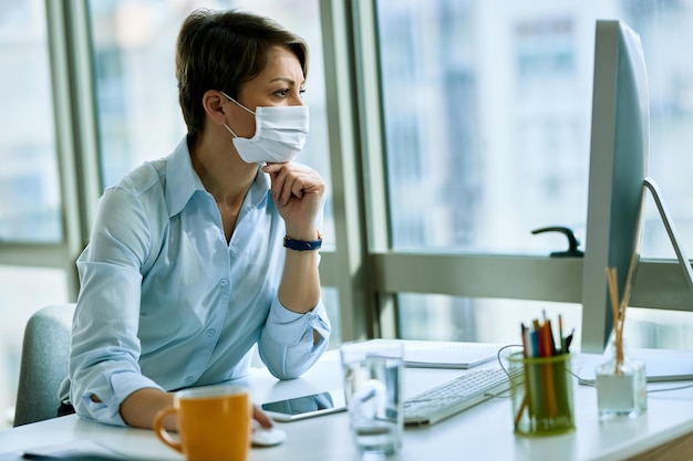 Donna d'affari che indossa una maschera facciale mentre legge un'e-mail sul PC desktop e lavora in ufficio