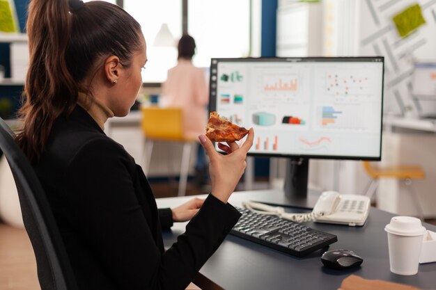 Donna d'affari che si gode l'ordine del pasto nell'ufficio dell'azienda durante la pausa pranzo da asporto lavorando ai grafici finanziari