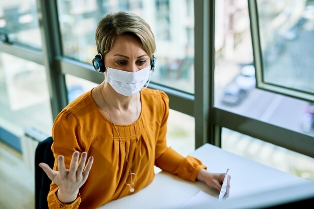 Donna d'affari con auricolare che comunica mentre lavora in ufficio e indossa una maschera facciale