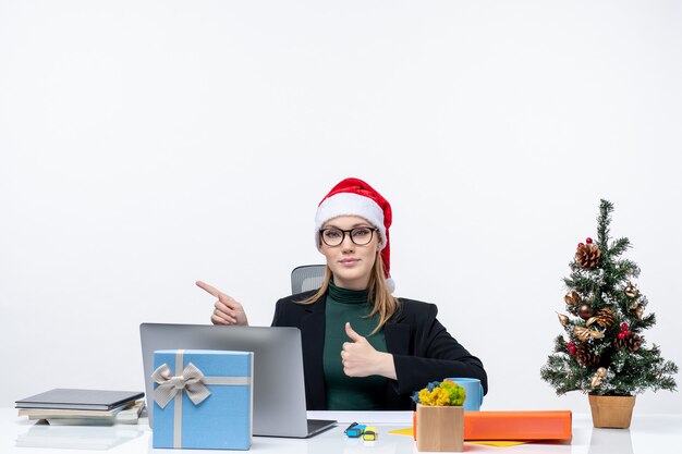 Donna d'affari con cappello di Babbo Natale seduto a un tavolo con un albero di Natale e un regalo