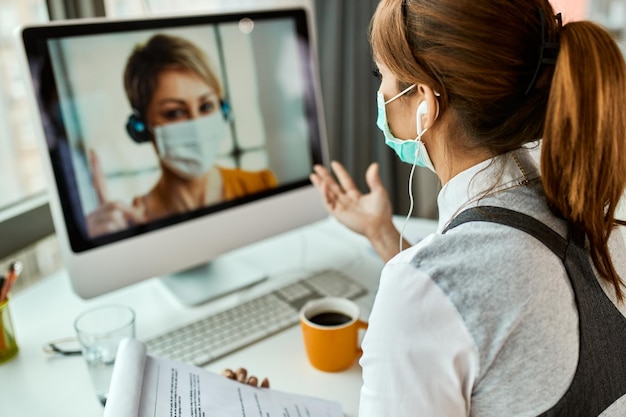 Donna d'affari con maschera facciale che comunica con il suo collega tramite videochiamata mentre lavora in ufficio durante la pandemia COVID19