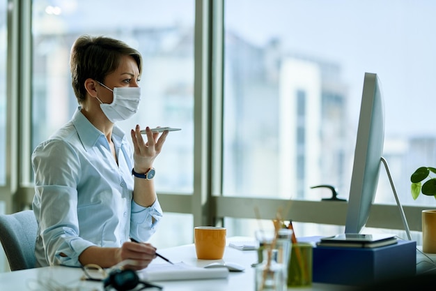 Donna d'affari con maschera facciale che lavora su un computer durante la registrazione di messaggi vocali su smartphone in ufficio