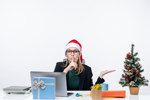 Donna d'affari con un cappello di Babbo Natale seduto a un tavolo con un albero di Natale e un regalo su di esso e indicando qualcosa sul lato sinistro qualcosa e facendo un gesto di silenzio in ufficio