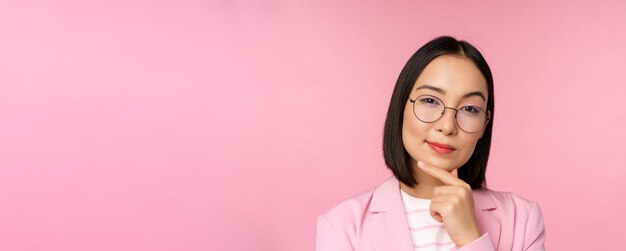 Donna d'affari coreana che pensa con gli occhiali che guarda premurosa alla macchina fotografica che prende una decisione in piedi su sfondo rosa