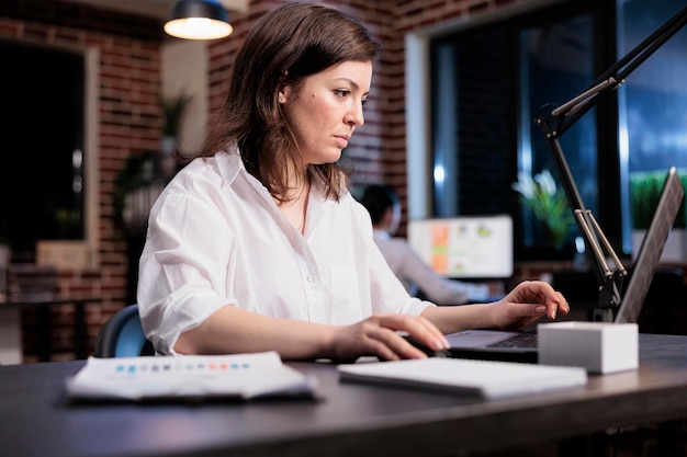 Donna d'affari di una società di marketing seduta nell'area di lavoro dell'ufficio durante la revisione della documentazione contabile. Project manager di avvio che utilizza il laptop per analizzare i grafici finanziari e promuovere il budget della campagna.