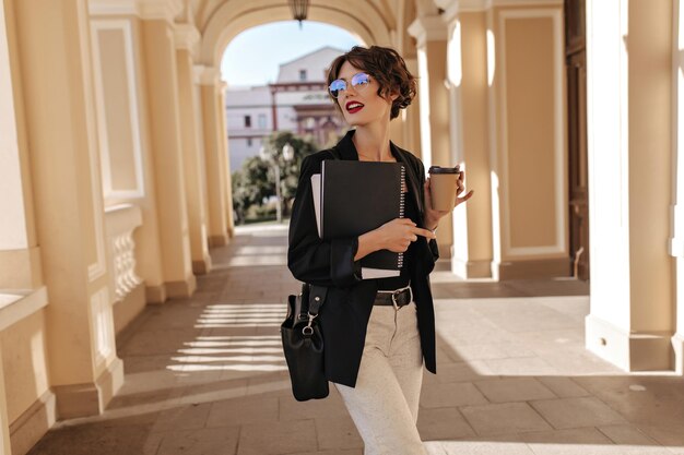 Donna d'affari in giacca scura e pantaloni bianchi in posa con una tazza di caffè all'esterno Ragazza alla moda con borsetta che tiene taccuino in strada