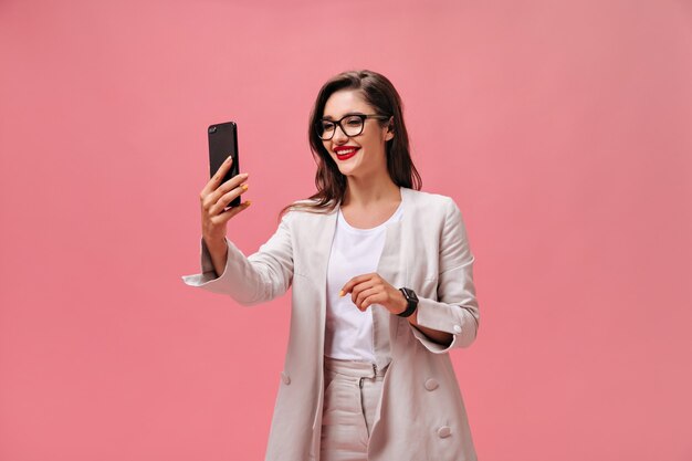 Donna d'affari in occhiali da vista e tuta prende selfie su sfondo rosa. Gioiosa ragazza affascinante con lunghi capelli scuri con rossetto rosso fa foto.