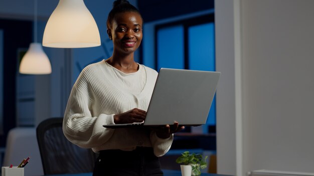 Donna d'affari nera che guarda la parte anteriore sorridente che tiene un laptop in piedi vicino alla scrivania in una società di start-up a tarda notte