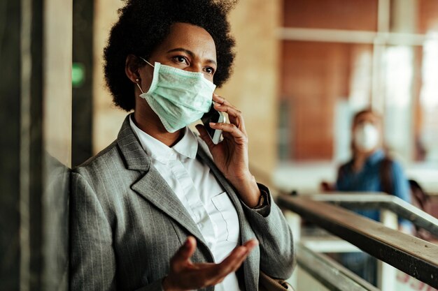 Donna d'affari nera che indossa una maschera protettiva durante la comunicazione sul telefono cellulare nel corridoio della stazione ferroviaria