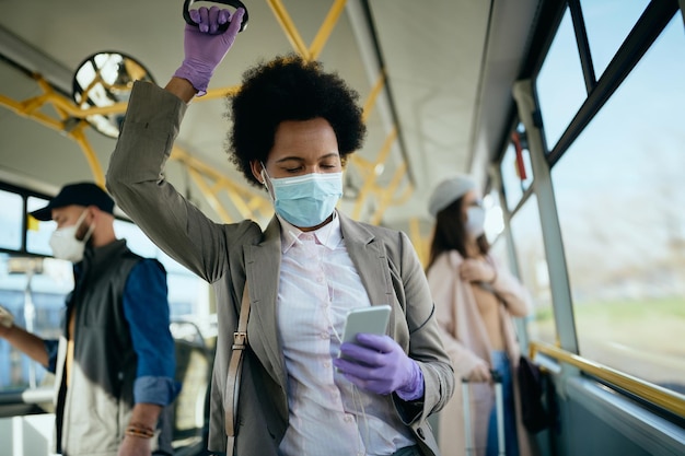 Donna d'affari nera che invia messaggi di testo sul cellulare e indossa una maschera durante il pendolarismo in autobus