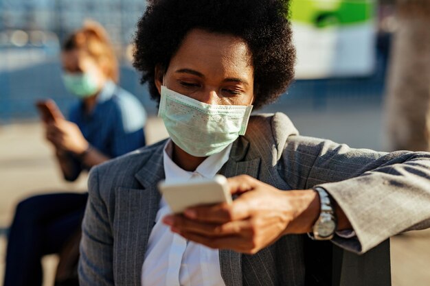 Donna d'affari nera con maschera protettiva sul viso tramite telefono cellulare e messaggi di testo per strada