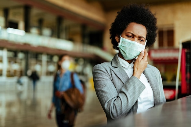 Donna d'affari nera dispiaciuta con maschera protettiva sul viso che parla al telefono cellulare al terminal dell'aeroporto