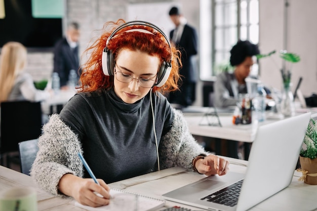 Donna d'affari rossa che usa il laptop e scrive note nel suo blocco note mentre ascolta musica in cuffia al lavoro