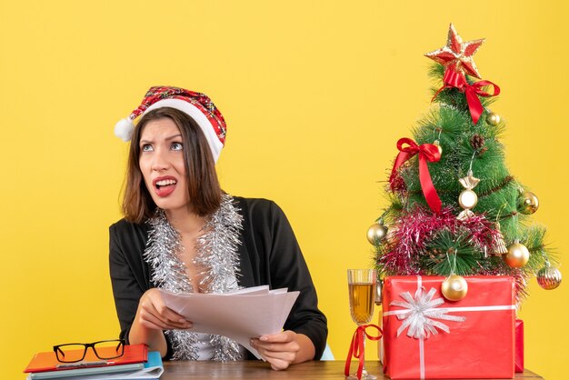 Donna d'affari scioccata in tuta con cappello di babbo natale e decorazioni di capodanno che tiene documenti e seduta a un tavolo con un albero di natale su di esso in ufficio