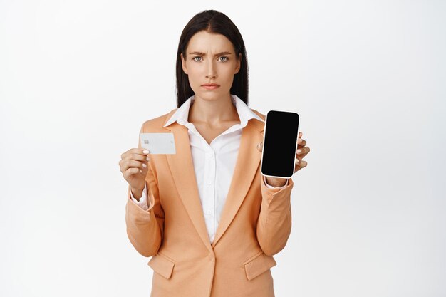 Donna d'affari sconvolta che mostra lo schermo vuoto del telefono cellulare e la carta di credito accigliata e che sembra delusa in piedi in tuta su sfondo bianco