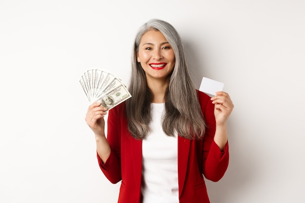 Donna d'affari senior asiatica di successo che mostra soldi in dollari e carta di plastica, sorride felice alla telecamera, indossa blazer rosso e trucco