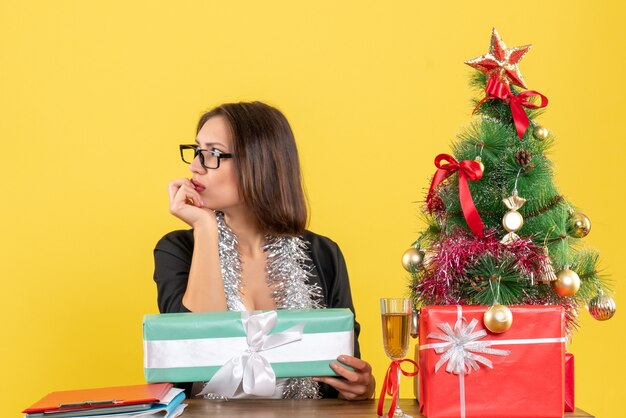 Donna d'affari sorpresa in vestito con gli occhiali in pensieri profondi che punta il suo regalo e seduto a un tavolo con un albero di Natale su di esso in ufficio