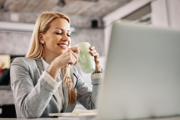 Donna d'affari sorridente che si prende una pausa e si gode una tazza di caffè mentre si siede alla sua scrivania in ufficio