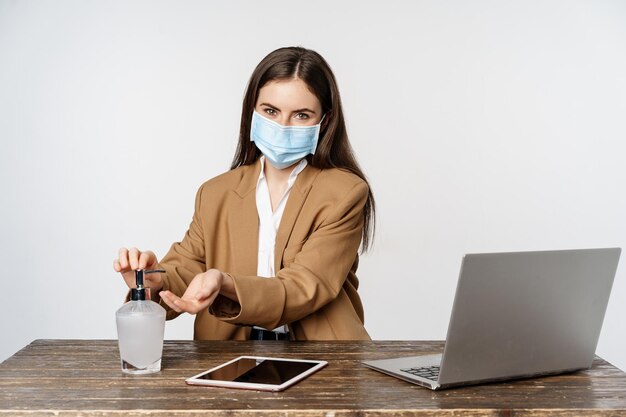 Donna d'affari sul posto di lavoro che indossa una maschera da covid-19, lavora in ufficio durante la pandemia, usa un disinfettante per le mani per pulire le mani, posa su sfondo bianco