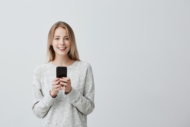 Donna dai capelli biondi felice positiva con ampio sorriso tramite cellulare, felice di ricevere un messaggio con una buona notizia, controllando il feed delle notizie sui suoi account di social network. Tecnologie e comunicazione moderne