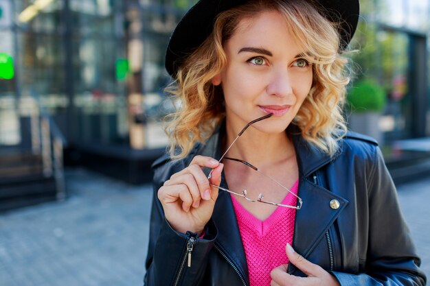 Donna dai capelli corti bionda pensierosa che cammina per le strade della grande città moderna. Vestito urbano alla moda. Occhiali da sole rosa insoliti.