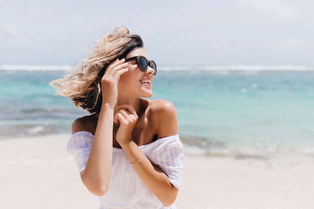 Donna dai capelli corti rilassata in posa sulla spiaggia. Colpo all'aperto di giovane signora allegra in occhiali da sole che gode della vacanza.