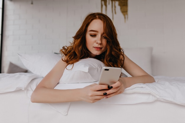 Donna dai capelli rossi interessata sdraiata a letto e guardando lo schermo del telefono. Modello femminile pigro con capelli rossi in posa in camera da letto.
