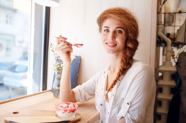 Donna dai capelli rossi sorridente che si siede in caffè e che mangia dessert