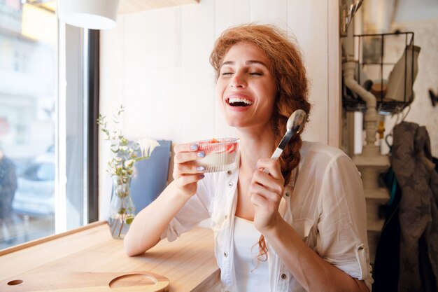 Donna dai capelli rossi sorridente che si siede in caffè e che mangia dessert