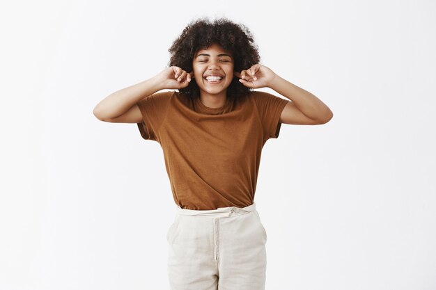 Donna dalla carnagione scura attraente incurante e felice con capelli ricci in vestito elegante, chiudendo gli occhi e sorridendo con espressione spensierata indifferente che copre le orecchie con le dita indice