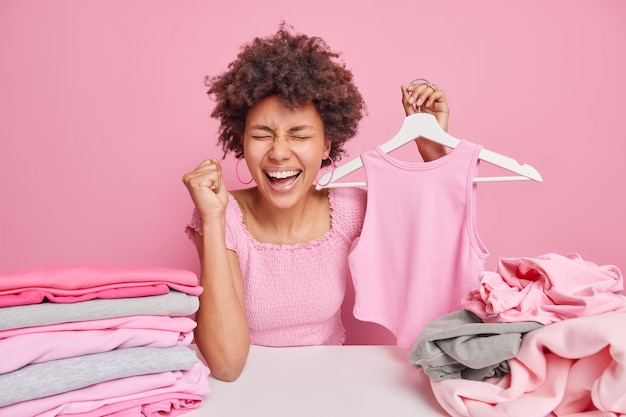 Donna dalla pelle scura felicissima con i capelli ricci stringe il pugno dalla felicità tiene l'abbigliamento sul gancio si siede al tavolo piega il bucato isolato sul muro rosa