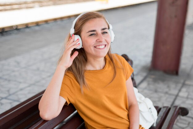 Donna del colpo metà che ascolta la musica sulla panchina nella stazione ferroviaria