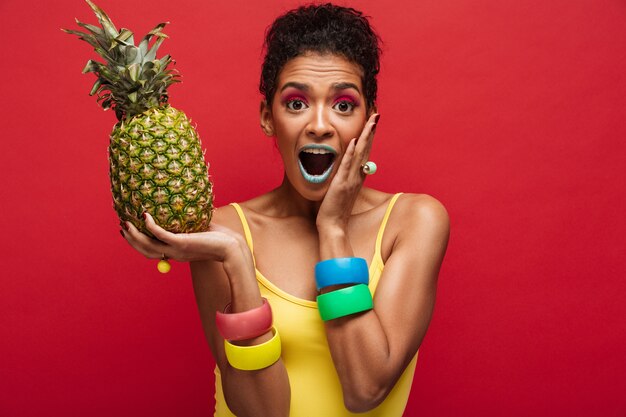 Donna del mulatto in attrezzatura variopinta che è eccitata mentre tenendo in mani l'ananas succoso fresco che gode della frutta isolata, sopra la parete rossa