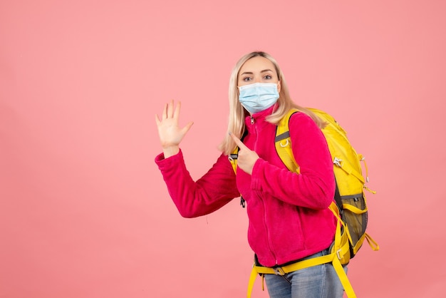 Donna del viaggiatore di vista frontale con lo zaino giallo che indossa la maschera che dà il cinque