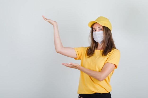 Donna delle consegne che mostra qualcosa con le mani in alto in t-shirt, pantaloni, berretto e maschera e sembra felice